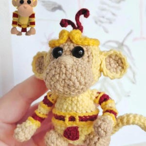 Puede incluir: Muñeco de mono de ganchillo hecho a mano en amarillo, beige y rojo. El mono tiene grandes ojos negros, una cara beige y orejas amarillas. Viste un atuendo amarillo con mangas a rayas rojas y amarillas y un cinturón rojo. Una imagen más pequeña del mono está en la esquina superior izquierda.