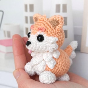 Puede incluir: Figura de perro corgi de ganchillo hecha a mano en melocotón, blanco y marrón. El perro tiene ojos negros grandes, una lengua rosa y un pecho blanco esponjoso. La figura se sostiene en una mano.