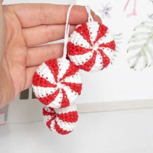 May include: Three crocheted peppermint candy ornaments in red and white. The ornaments are hanging from a string and are held in a hand.
