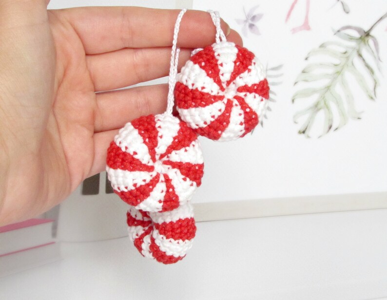 May include: Three crocheted peppermint candy ornaments in red and white. The ornaments are hanging from a string and are held in a hand.