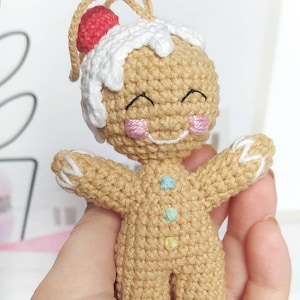 May include: A crocheted gingerbread man ornament with a white icing top and red candy. The gingerbread man has a smiling face with pink cheeks and is decorated with colorful buttons.