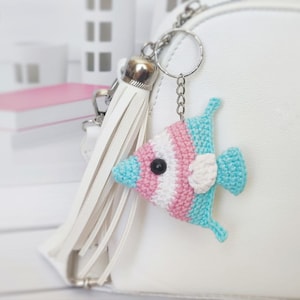 Op de afbeelding: Een witte gehaakte vis sleutelhanger met roze, blauwe en witte strepen. De vis heeft een zwart oog en een witte vin. De sleutelhanger is bevestigd aan een zilveren ketting en een witte kwast.