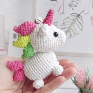 May include: A white crocheted unicorn with a pink horn, mane, and tail. The unicorn has black eyes and a silver ring attached to its head. The mane is a rainbow of colours, including pink, yellow, and green. The unicorn is a handmade item.