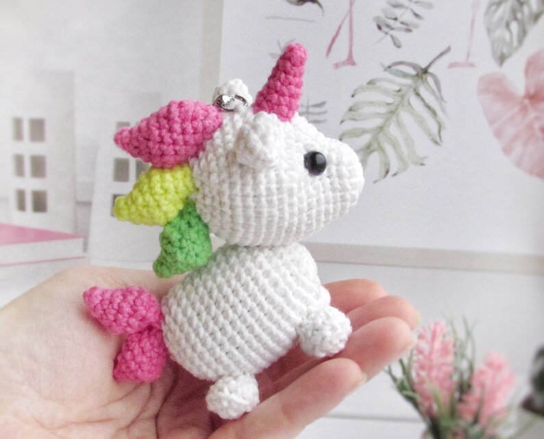 May include: A white crocheted unicorn with a pink horn, mane, and tail. The unicorn has black eyes and a silver ring attached to its head. The mane is a rainbow of colours, including pink, yellow, and green. The unicorn is a handmade item.