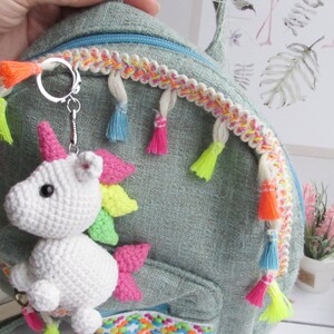 May include: A white crocheted unicorn keyring with a pink horn and colourful mane is attached to a teal backpack. The backpack has a decorative trim with tassels in various bright colours. The keyring is a whimsical accessory.