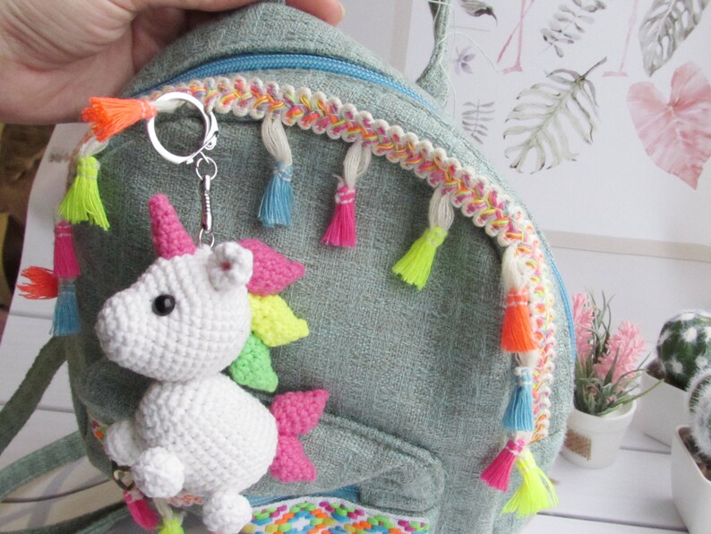 May include: A white crocheted unicorn keyring with a pink horn and colourful mane is attached to a teal backpack. The backpack has a decorative trim with tassels in various bright colours. The keyring is a whimsical accessory.