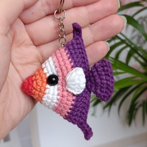 Puede incluir: Un llavero de pescado de crochet con un esquema de color morado, rosa, blanco y naranja. El pez tiene un ojo negro y una barriga blanca.