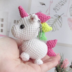 May include: A white crocheted unicorn keyring with a pink horn, rainbow mane, and tail. The unicorn has black eyes and a silver keyring. The handmade unicorn is a whimsical accessory.