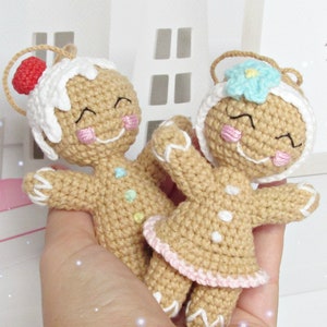 May include: Two crocheted gingerbread person ornaments. One is wearing a white frosting hat with a red candy on top. The other is wearing a blue flower on top of its head. Both have pink cheeks and smiling faces.