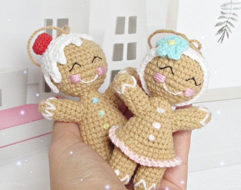 May include: Two crocheted gingerbread people ornaments. One is wearing a white frosting hat with a red candy on top. The other is wearing a blue flower on top of its head. Both have pink cheeks and smiling faces.