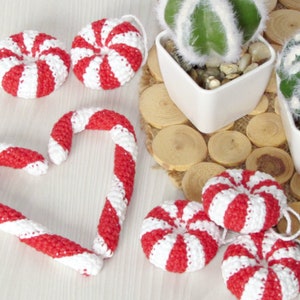 May include: A set of crocheted candy cane ornaments in red and white, including a heart-shaped candy cane and round candy canes. The ornaments are displayed on a white surface with a background of potted cacti.