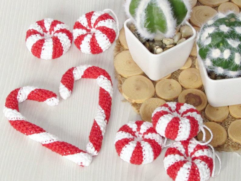 May include: A set of crocheted candy cane ornaments in red and white, including a heart-shaped candy cane and round candy canes. The ornaments are displayed on a white surface with a background of potted cacti.