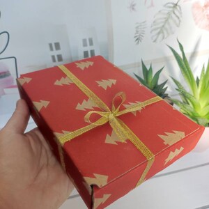 May include: A red gift box with a gold ribbon and a pattern of small Christmas trees. The box is wrapped in red paper with a gold ribbon tied in a bow.