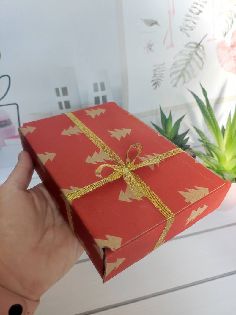 May include: A red gift box with a gold ribbon and a pattern of small Christmas trees. The box is wrapped in red paper with a gold ribbon tied in a bow.