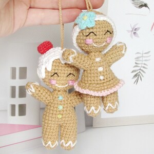 May include: Two crocheted gingerbread people ornaments, one with a pink and white icing design and the other with a blue and white icing design. Both have smiling faces and are holding their arms out.