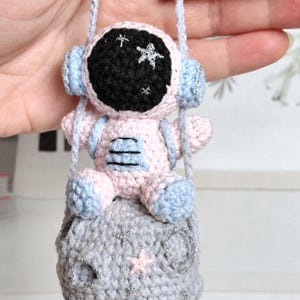 May include: A crocheted astronaut doll in a pink and blue spacesuit, seated on a gray crocheted moon. The astronaut's helmet is black with silver star accents. The moon has silver star details and a pink star. A light blue cord is attached.