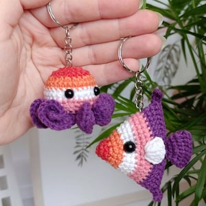 Könnte beinhalten: Zwei gehäkelte Schlüsselanhänger, einer in Form eines Kraken und der andere in Form eines Fisches, beide mit einem Farbschema der lesbischen Pride-Flagge. Der Krake ist lila, weiß, orange und rosa. Der Fisch ist lila, weiß, orange und rosa. Beide sind an silbernen Schlüsselringen befestigt.