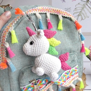 May include: A white crocheted unicorn keyring with a rainbow mane and pink horn is attached to a small, pale green backpack. The backpack has colourful trim and tassels. The keyring is a whimsical accessory.