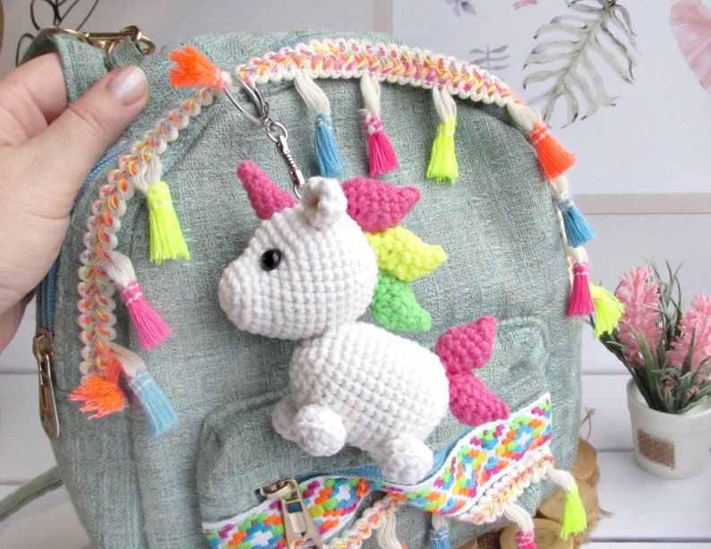 May include: A white crocheted unicorn keyring with a rainbow mane and pink horn is attached to a small, pale green backpack. The backpack has colourful trim and tassels. The keyring is a whimsical accessory.