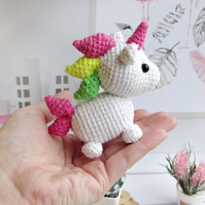 May include: A small, white crocheted unicorn with a pink horn, mane, and tail. The unicorn has black eyes and is posed as if walking. The mane is a rainbow of colours, including pink, yellow, and green. The unicorn is a handmade item.