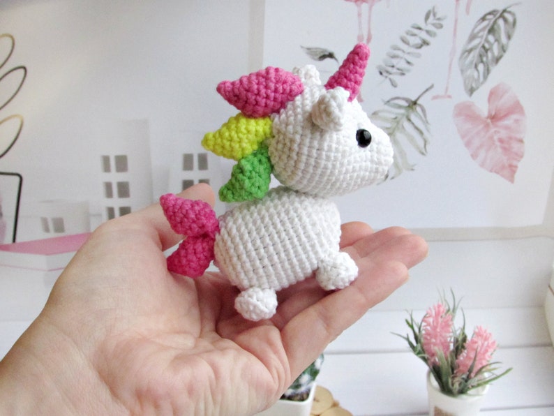 May include: A small, white crocheted unicorn with a pink horn, mane, and tail. The unicorn has black eyes and is posed as if walking. The mane is a rainbow of colours, including pink, yellow, and green. The unicorn is a handmade item.