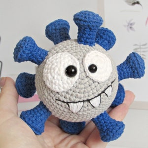 May include: A gray crocheted plush toy shaped like a virus with blue spikes and a toothy grin. The toy has large white eyes with black pupils.