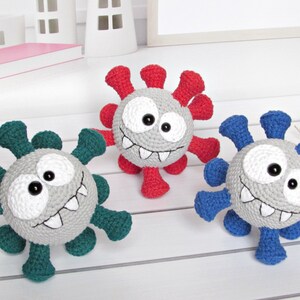 May include: Three crocheted plush toys shaped like viruses. The viruses are gray with white eyes and teeth. The viruses have different colored spikes: red, blue, and green.