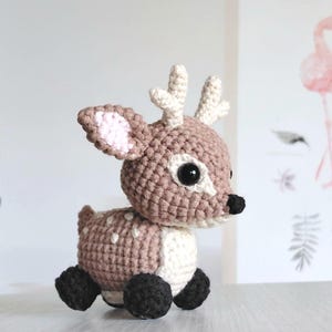 May include: A handmade crocheted deer toy in brown, cream, and black. The deer features large black eyes, cream antlers, and a white belly with brown spots. The ears have a pink interior. The toy is sitting on a surface.