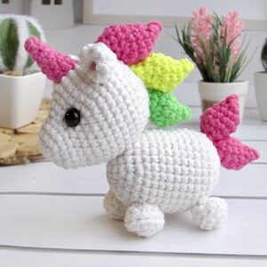 May include: A white crocheted unicorn with a pink horn, pink and yellow mane, and pink tail. The handmade stuffed animal has black eyes and is posed on a white surface. The unicorn is a whimsical, handmade toy.
