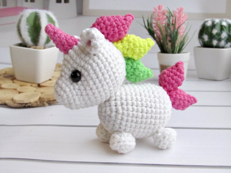 May include: A white crocheted unicorn with a pink horn, pink and yellow mane, and pink tail. The handmade stuffed animal has black eyes and is posed on a white surface. The unicorn is a whimsical, handmade toy.