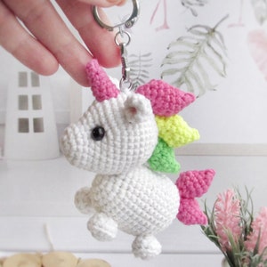 May include: A white crocheted unicorn keyring with a rainbow mane and tail. The unicorn has pink horns and black eyes. The keyring is attached to a silver metal clasp.