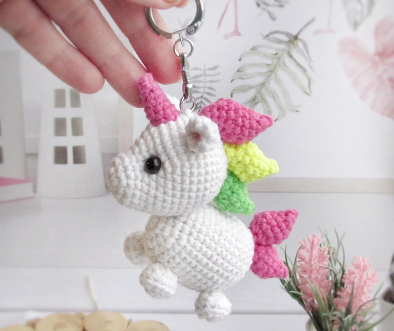 May include: A white crocheted unicorn keyring with a rainbow mane and tail. The unicorn has pink horns and black eyes. The keyring is attached to a silver metal clasp.