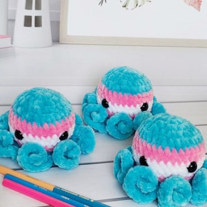 May include: Three crocheted octopus plushies in blue, pink, and white. The plushies have black eyes and are sitting on a white surface.