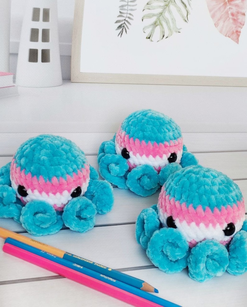 May include: Three crocheted octopus plushies in blue, pink, and white. The plushies have black eyes and are sitting on a white surface.