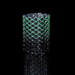 May include: A cylindrical object featuring a green, diamond-shaped lattice pattern that transitions into a solid black base. The structure is open at the top, revealing a dark interior, and displays a repeating geometric mesh design throughout.