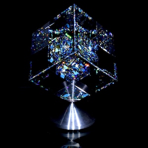 May include: A modern, geometric sculpture featuring a mirrored cube filled with colorful, fragmented glass. The cube sits atop a silver, conical base, reflecting light and creating a dazzling display. The artwork is set against a black background.