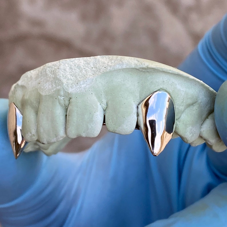 Puede incluir: Una imagen de primer plano muestra un conjunto de dientes de vampiro plateados, tambi&eacute;n conocidos como colmillos, unidos a un molde de boca. Los dientes est&aacute;n siendo sostenidos por una mano enguantada.