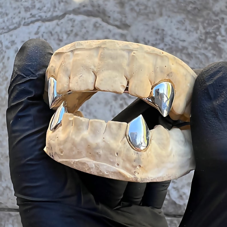 Puede incluir: Una imagen de primer plano de un conjunto de dientes de vampiro plateados, tambi&eacute;n conocidos como colmillos. Los dientes est&aacute;n hechos de metal y est&aacute;n dise&ntilde;ados para ajustarse sobre los dientes existentes. Son un accesorio popular para los disfraces de Halloween y otros eventos.