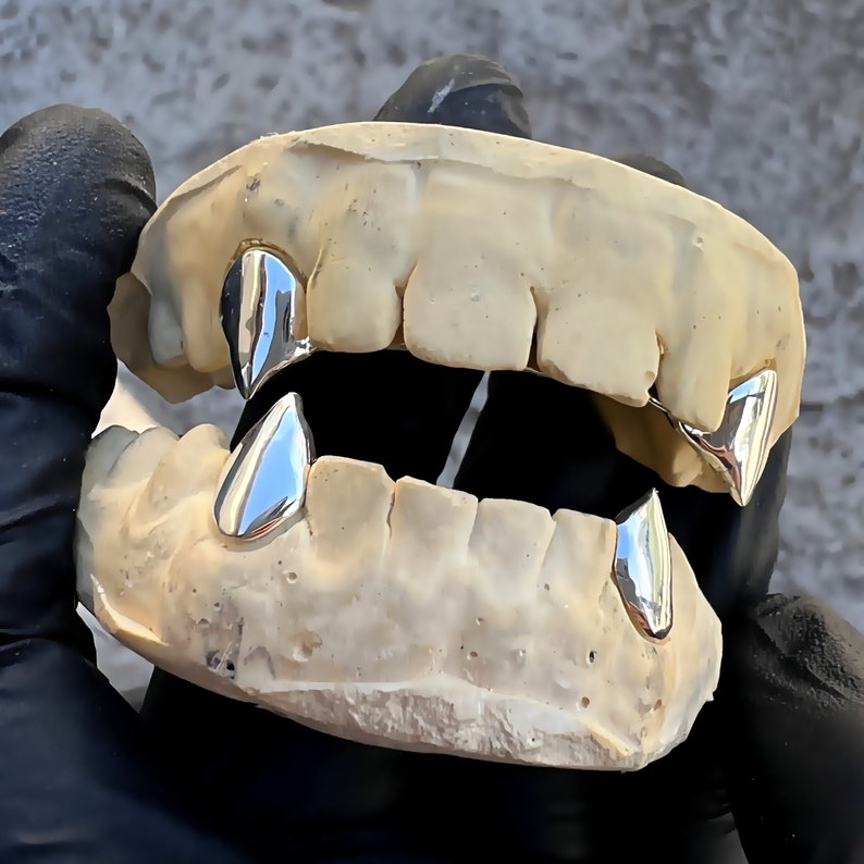 Puede incluir: Primer plano de un juego de dientes de oro, tambi&eacute;n conocidos como grills, con un dise&ntilde;o puntiagudo en forma de colmillos. Las grills est&aacute;n hechas de metal y est&aacute;n dise&ntilde;adas para ajustarse sobre los dientes del usuario.