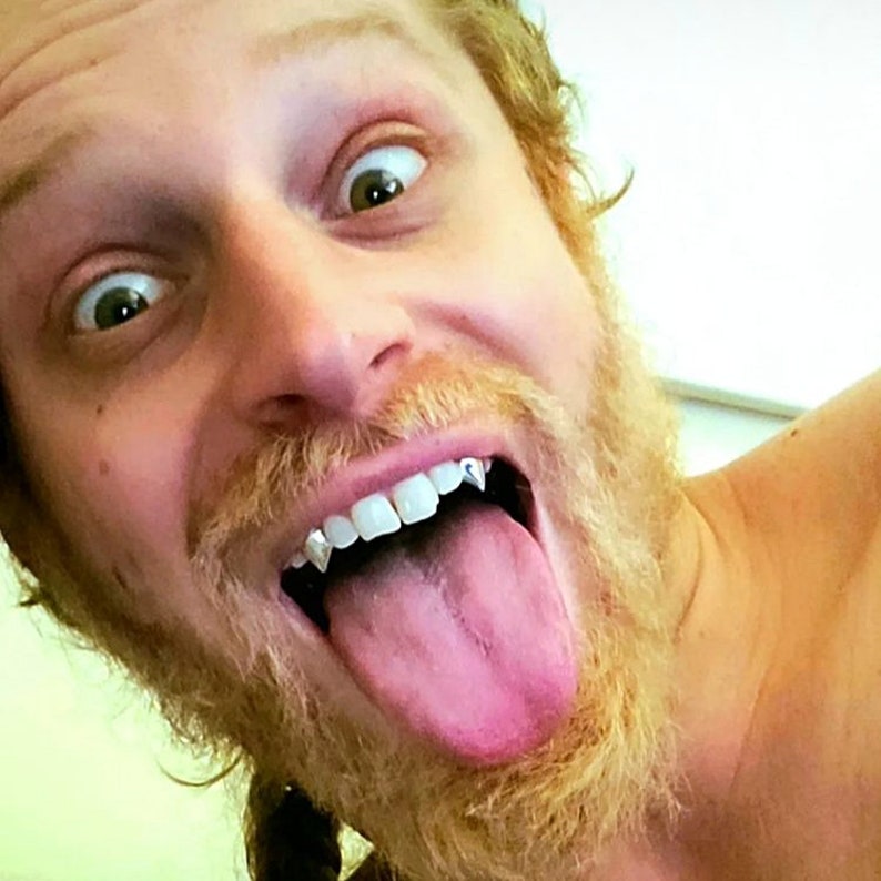 Puede incluir: Primer plano de un hombre con una barba larga y casta&ntilde;a clara, sacando la lengua. Lleva un juego de colmillos de vampiro de oro.