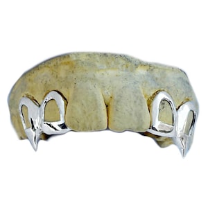 May include: A set of silver-colored fang-shaped grillz, designed to fit over the upper teeth. The grillz are highly polished and reflect light, creating a shiny appearance. The teeth mold is a light beige color.