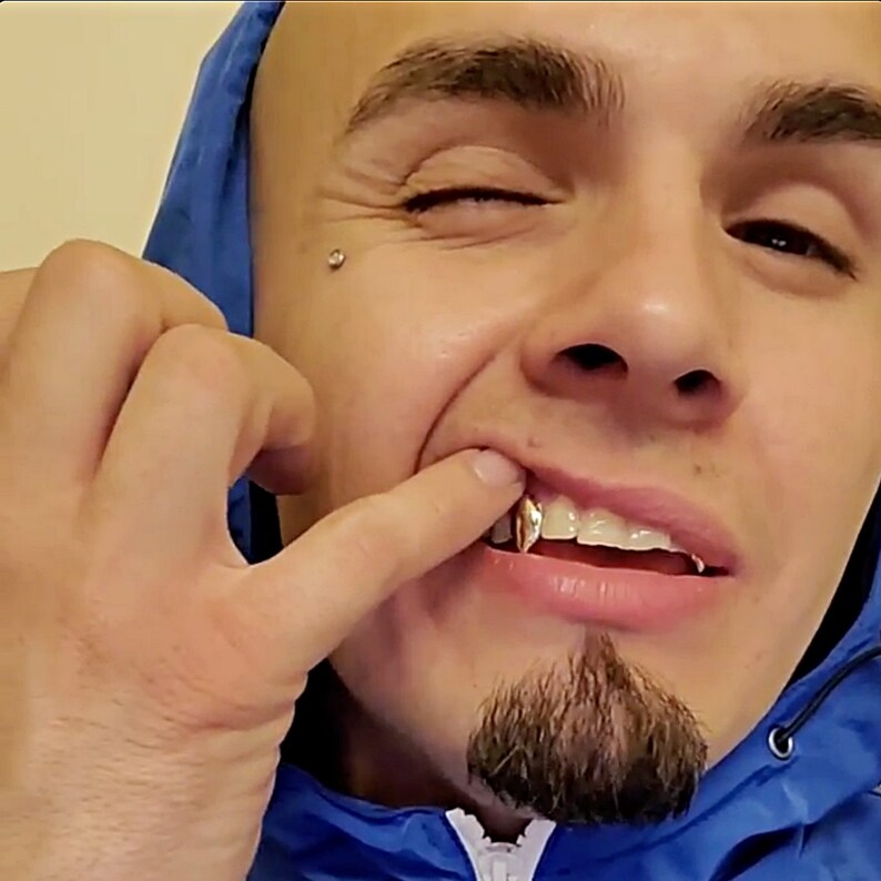 May include: A close-up of a person's mouth with gold teeth. The person is wearing a blue hooded jacket and has a small stud earring in their left ear.