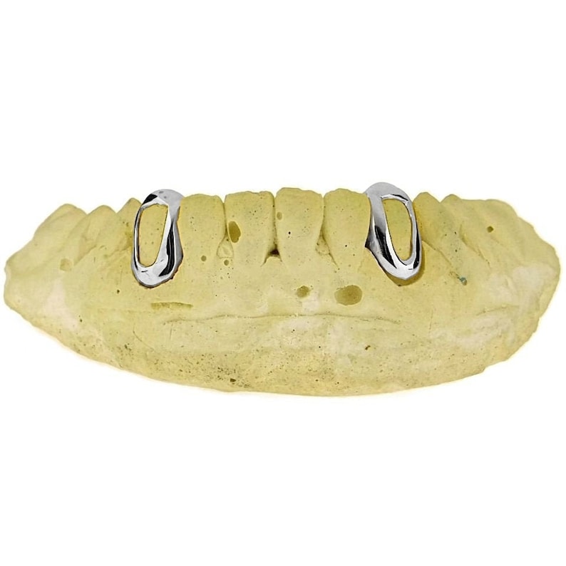 May include: Two silver-colored dental caps, also known as grills, are shown on a yellow dental mold. The caps are oval-shaped and have a smooth, polished finish.