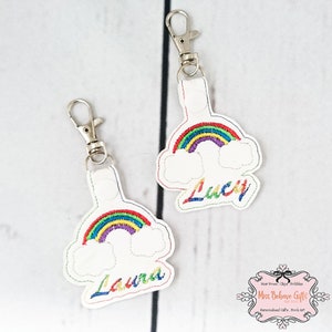 Könnte beinhalten: Zwei weiße Stoffschlüsselanhänger mit Regenbogen- und Wolken-Stickerei. Die Schlüsselanhänger sind mit den Namen "Laura" und "Lucy" bestickt.