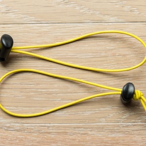 Puede incluir: Dos cordones elásticos amarillos con botones de plástico negro. Los cordones están en bucle y atados juntos.