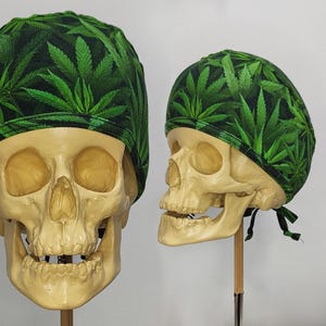 May include: Two skull models wearing green and black patterned caps. The caps feature a repeating pattern of green cannabis leaves. The skulls are positioned on wooden stands against a white background.