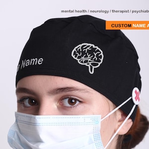 May include: Black scrub cap with a white embroidered brain design and the text "Name". The cap has a button with a red cross for mask straps. Text at the top says "CUSTOM NAME AVAILABLE".