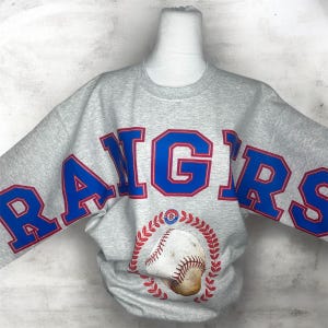 May include: A heather gray crewneck sweatshirt featuring the word RANGERS in large blue block letters with red outlines across the chest. Below the text is a graphic of a baseball surrounded by a red circular laurel wreath design.