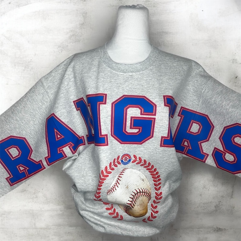 May include: A heather gray crewneck sweatshirt featuring the word RANGERS in large blue block letters with red outlines across the chest. Below the text is a graphic of a baseball surrounded by a red circular laurel wreath design.