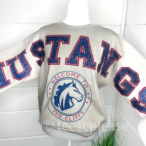 May include: A beige crewneck sweatshirt featuring the word MUSTANGS in large, distressed navy blue letters with red outlines. A circular graphic on the chest displays a horse head profile with the text WELCOME TO THE CLIFFS in navy blue.